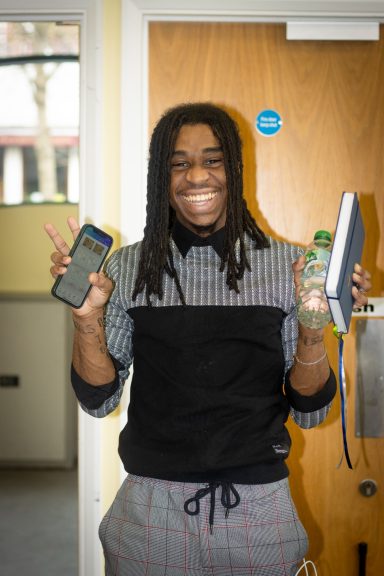 Person smiling and holding a phone, a drink, and a notebook in a casual setting.