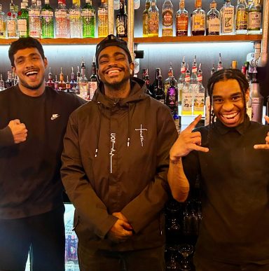 Three smiling young men stand in front of a bar with various bottles behind them.