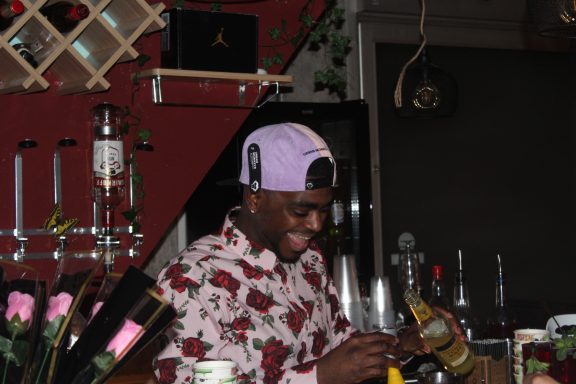 A young man in a floral shirt smiles while preparing drinks at a bar.